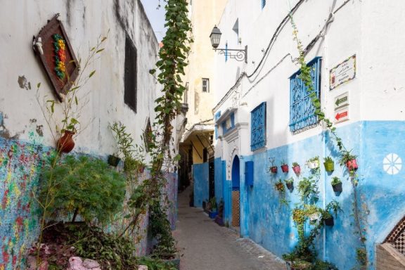 Rue étroite avec des murs bleus, plantes grimpantes et décorations colorées.